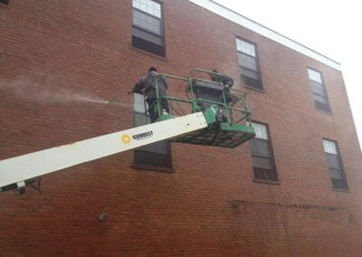 Two workers from a pressure washing company are pressure cleaning the exterior brick wall of a building from a raised platform lift.
