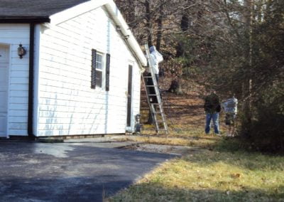 Two people near a house; one on a ladder working on the roof, the other standing nearby holding equipment from a pressure washing company.