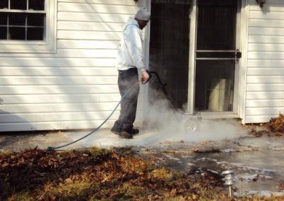 A person from a Pressure Washing Company is using a pressure washer on a patio outside a white house with leaves scattered on the ground.