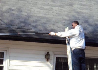 A person on a ladder in a white hoodie and dark pants is carefully inspecting or cleaning the roof of a house, ensuring it stays pristine with the expert touch of pressure cleaning services.