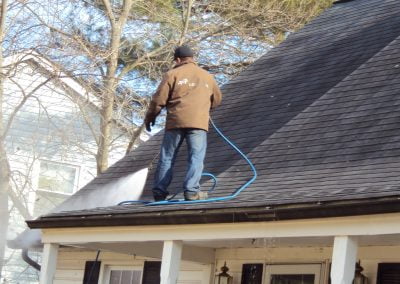 A person standing on a roof power-washing shingles with a hose. Trees and a house are visible in the background. It looks like professional pressure cleaning services by a reputable Pressure Washing Company.