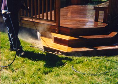 A person using a pressure washer to clean a wooden deck in a backyard with lush green grass and a house in the background, showcasing the great results of professional pressure cleaning services.