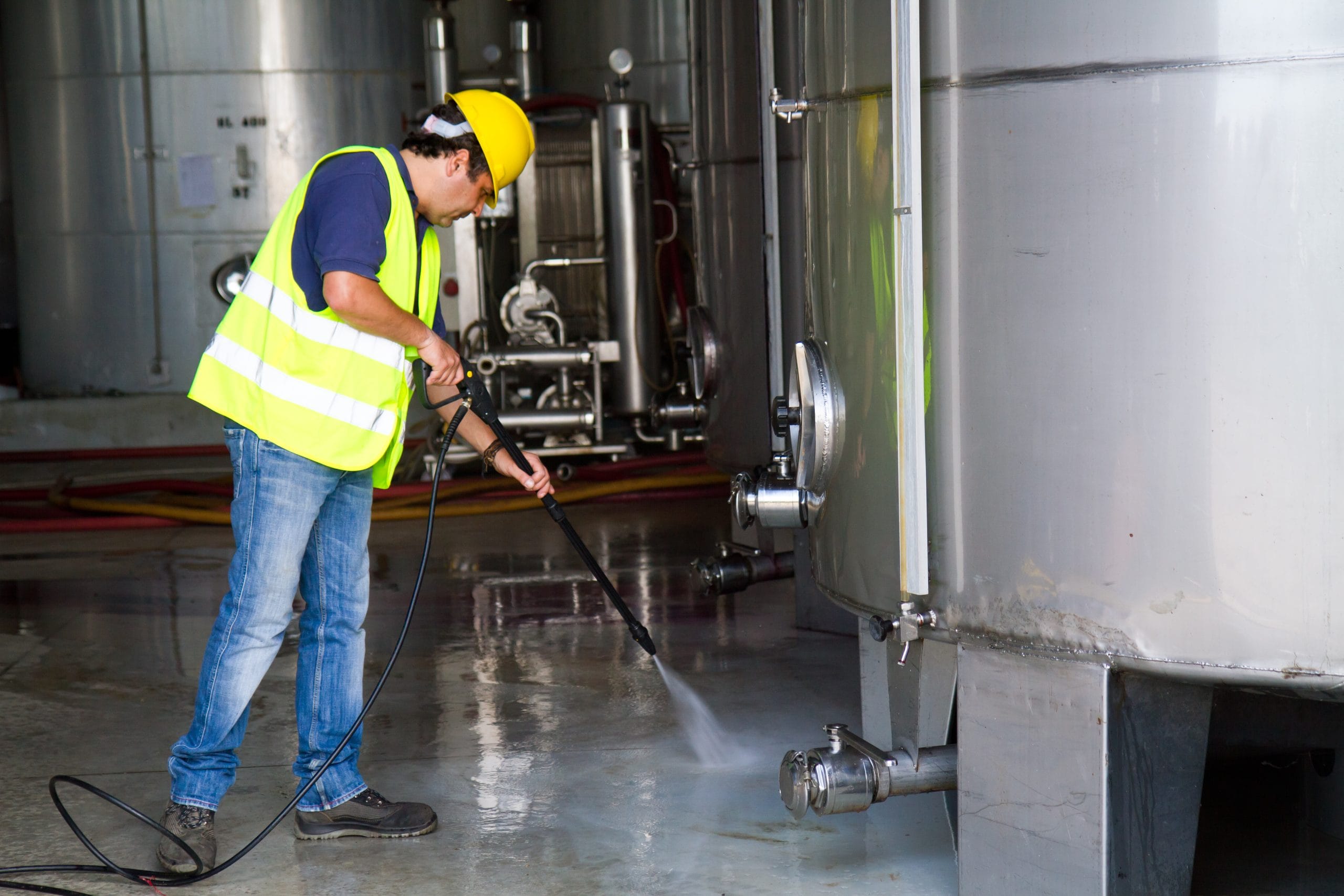 washing industrial washing Worker in a yellow vest and hard hat uses a pressure washer on a large metal tank in an industrial setting.