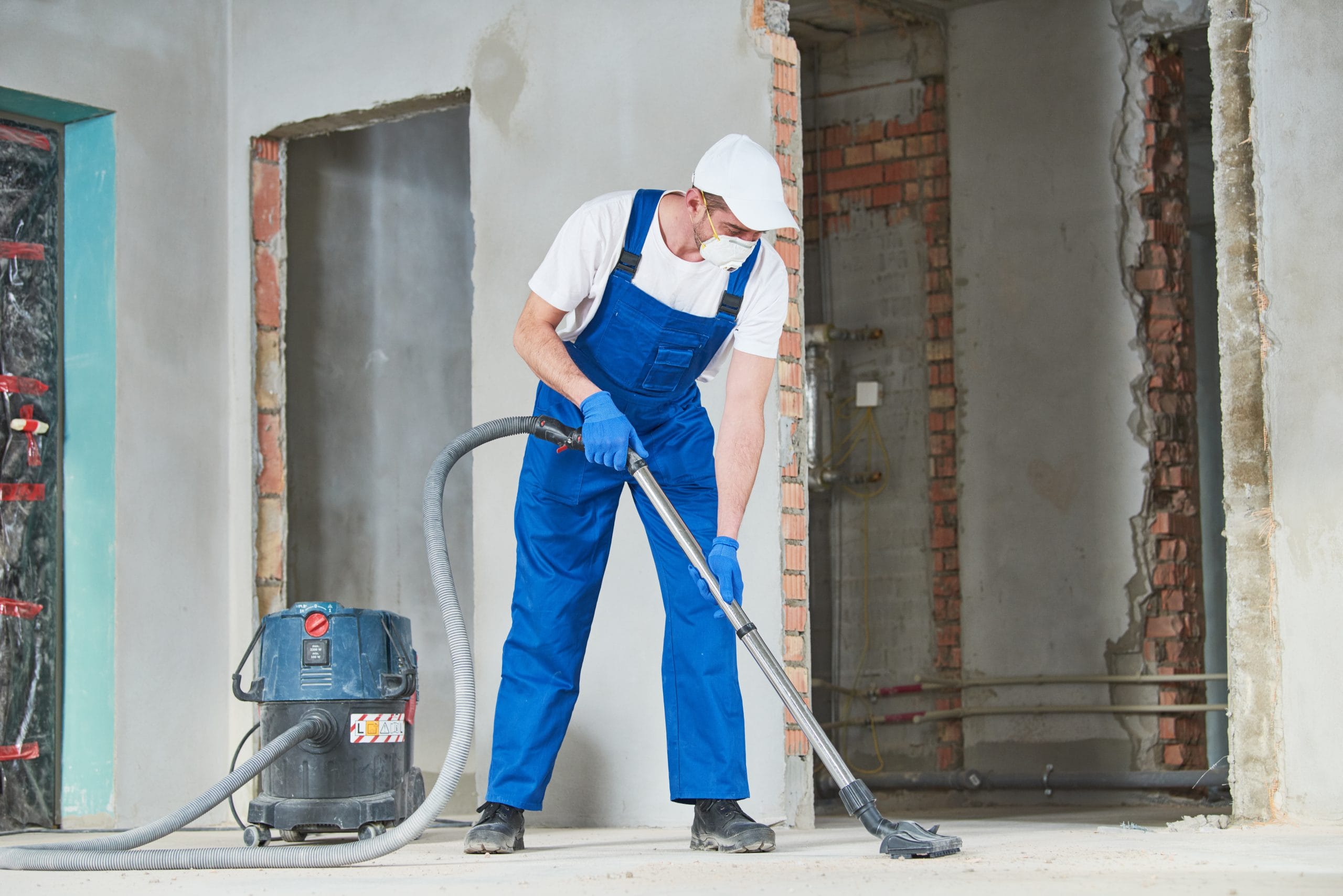 Worker in blue overalls and mask vacuuming debris at a construction site with exposed brick and unfinished walls.